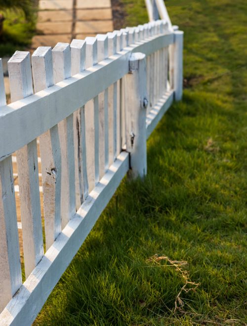 Close-up view of a white wooden fence installed against the sunlight on the lawn near the concrete stairs with concrete slabs as a pathway in the Thai countryside.
